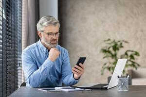 Mature businessman looking at smartphone while working on laptop in a modern office, emphasizing focus and concentration. photo