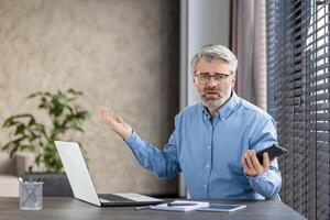 Confused businessman working from home with a laptop and smartphone, expressing frustration. He's seated at a desk with notes and office supplies. photo