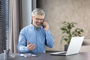 Smiling mature businessman communicating on phone while working on laptop at desk in a modern office with natural light and greenery in the background. photo