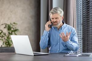 Middle-aged man in office setting showing frustration during a phone call. He is sitting at a desk with a laptop and notepad, indicating a professional work environment. photo