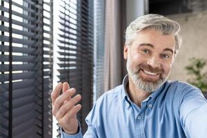 Smiling man with grey hair and beard having a call in bright office with blinds in the background. photo