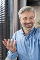 Smiling mature man in blue shirt having call using phone from office or home, engaging in conversation, looking at camera, and showing positive emotions photo