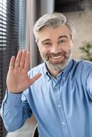 A cheerful mature office worker waves at the camera, greeting on a call. Wearing a blue shirt, he presents a warm and friendly demeanor in a professional office setting photo
