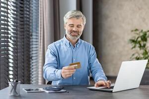 Mature man smiling and using a credit card for online payment on his laptop while sitting at a home office desk. photo