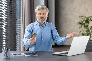 Senior man looking frustrated while holding a credit card in one hand and gesturing with the other hand at home. He is sitting in front of a laptop, facing an online payment issue. photo