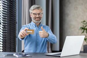 Smiling man holding credit card and showing thumbs up gesture at desk with laptop, smartphone, and notebook. Concept of online shopping, ecommerce, approval, and successful transaction photo