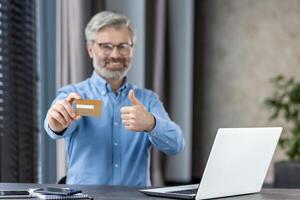 Smiling office worker in his 40s showing a credit card and giving a thumbs up gesture while working on a laptop. Represents satisfaction, approval, and success in business or online transactions. photo
