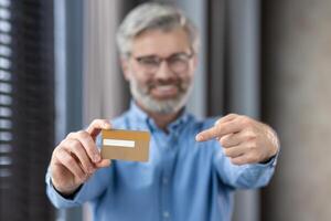 Middle-aged man with beard and glasses smiling, holding and pointing at a credit card, indoors photo