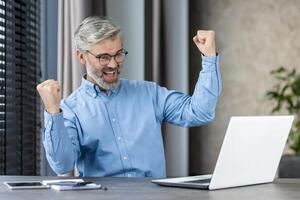 Happy businessman celebrating success while working on laptop in a modern office setting. photo