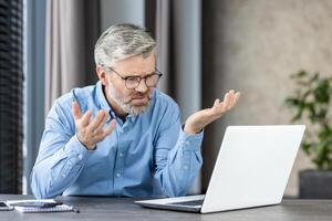 Senior man in a blue shirt expressing frustration while working on a laptop at home. The man appears to be looking for a solution or dealing with a technical problem. photo