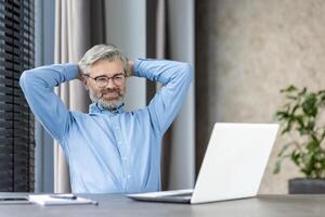 Mature businessman enjoying a break at home office desk, smiling, and looking relaxed with hands behind his head. photo