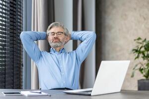 Mature businessman with glasses and beard taking a moment to relax at office desk. Hands behind head, laptop in front, serene expression. Concept of rest, leisure, workplace, and stress relief. photo