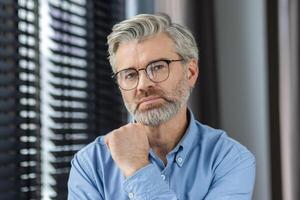 Confident mature man with glasses in a blue shirt posing indoors by window blinds, showcasing a thoughtful and reflective expression. photo