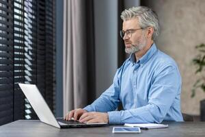 Mature man in glasses and blue shirt working on a laptop in a well-lit office space. He appears concentrated and professional, embodying productivity and efficiency in a modern work environment. photo
