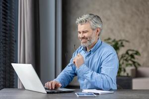 Senior man working on laptop, smiling while seated in a modern office. The man is wearing a blue shirt and appears engaged in his work. photo