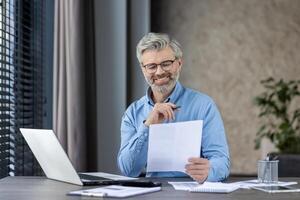 A smiling middle-aged businessman with glasses and beard reviews documents at his office desk. He is working on a laptop and appears content in a modern workspace. photo
