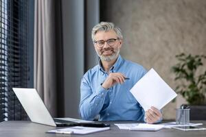 Mature businessman working at desk with laptop and documents in modern office, smiling confidently. Professional workspace with natural light. Successful and productive work environment photo