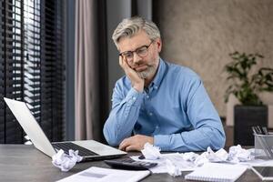 Senior man feeling stressed while working on a laptop at a desk cluttered with crumpled papers, indicating frustration at work. photo