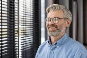 Mature man with glasses smiling while looking out window with blinds in contemporary office environment. Natural light illuminating cheerful expression capturing professional yet relaxed demeanor photo