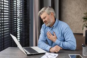 Elderly man touching chest in discomfort while sitting at desk with laptop, indicating possible heart problem or stress related illness in office environment photo