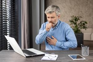 Middle-aged man working from home is seen coughing and holding his chest while seated at a desk with a laptop. The image captures the concept of health issues, remote work challenges, and stress. photo