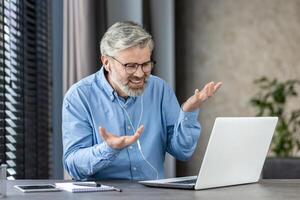 Senior man using a laptop and headset for a call at home. He is sitting at a desk and appears to be engaged in the conversation. photo