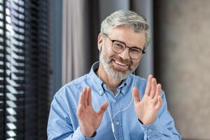 Senior man in an office setting wearing headphones, smiling and waving during a call. The man appears to be calm and happy as he engages in a remote work meeting. photo