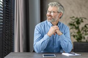 Thoughtful mature office worker sitting at his desk, looking out the window and contemplating. He is wearing a blue shirt and glasses, with a tablet and notebook on the table in a modern office photo