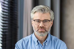 Close-up portrait of a mature man with glasses and beard wearing a blue shirt. Indoors with a blurred background. photo