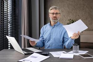 Professional businessman sitting at desk holding documents, displaying concern while working on laptop in modern office. Concept of work stress, business finance, office environment photo
