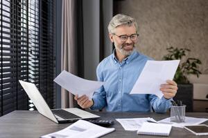Confident businessman smiling while reviewing documents at his desk with a laptop in a modern office. Professional setting with natural light and organized workspace. photo