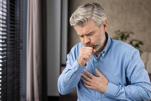 Elderly man with grey hair and beard coughing and holding chest with painful expression. Health concept of illness, bronchitis, lung disease, or flu. Indoors with window and blurred background. photo