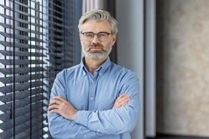 Portrait of caucasian businessman in eyeglasses standing with folded hands on apartment background. Successful aged male feeling calm and confident about future of company and economical stability. photo
