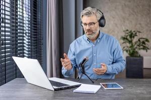 Focused aged male in headphones sitting by working table with laptop and professional microphone indoors. Busy bearded man recording podcast lecture while using modern technology for best acoustic. photo