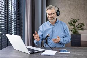 A gray-haired senior man is sitting at the table at home, wearing headphones in front of a microphone and chatting online via call, recording a webinar. photo