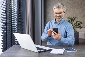 Satisfied middle aged freelancer using mobile phone by desktop with computer next to window with jalousie. Smiling relaxed worker reading pleasant text message during busy day at home workspace. photo