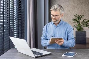 Thoughtful mature male with grey beard looking at portable computer screen while taking notes in domestic workplace. Attentive employee watching training webinar and making summary with main theses. photo