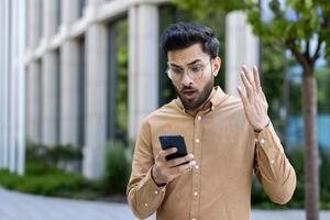 Man displaying shocked expression while looking at smartphone outside. Wearing glasses and brown shirt, reacting to unexpected news or message. Urban background includes modern building and greenery. photo