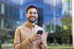 Confident young man with glasses holds smartphone while standing outside contemporary office. He is smiling at camera, suggesting positive communication and connectivity. photo