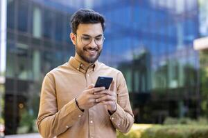 Smiling young man holding smartphone outdoors in urban setting. Represents connection, technology, communication, displaying positive feeling while interacting with phone near modern office building. photo
