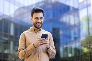 Young man smiling while using smartphone outside modern glass office building, symbolizing urban lifestyle, connectivity, and communication. photo