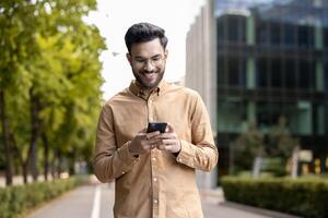 Smiling man wearing glasses and casual clothes using smartphone outdoors. Enjoying technology in urban setting with modern buildings. Capturing moment connectivity and communication through phone. photo