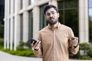 Man with glasses standing outside, holding phone and credit card, showing confused expression. Concept of financial uncertainty, technology frustration, decision making, or communication issues. photo