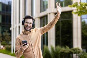Cheerful man wearing headphones smiling while holding phone and waving outside modern building. Person enjoying music and technology in casual setting, conveying happiness and connection in outdoor photo