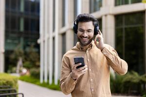 Smiling man in casual attire listens to music with headphones using phone, enjoying a pleasant walk outside modern office building. Captures essence of leisure, technology interaction photo