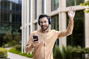 Smiling man wearing headphones uses smartphone enjoying music outdoors. Standing with hand raised in excitement, expressing happiness and enjoyment in sunny urban setting. photo