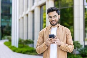 Smiling young man wearing glasses using smartphone outside modern office building. Casually dressed man engaging with social media or business apps phone, bright environment representing technology photo