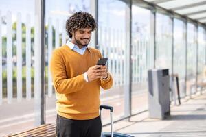 Man holding phone while standing at public transport stop. Passenger buys tickets remotely online, smiling happily, waiting for bus or tram. photo