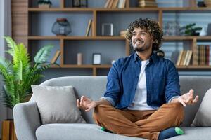 Joyful man at home resting and relaxing meditating in lotus pose sitting on sofa in living room of apartment, smiling contentedly looking out window. photo