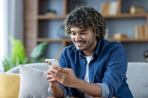 Young joyful man uses phone while sitting at home on sofa in living room of apartment, smiles happily, browses social media, and types text message. photo
